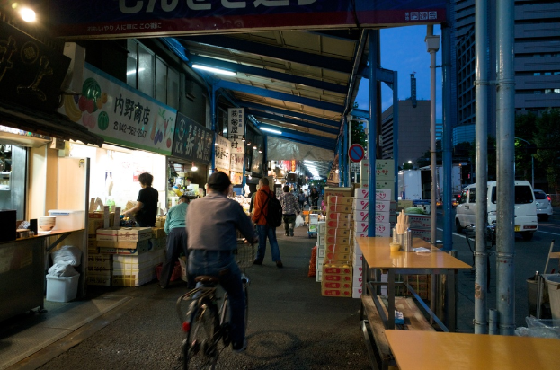 Ein Gehweg mit Geschäften auf der linken Seite und Kisten, einem Tisch, einer Straße mit Autos, Bäumen und einem Gebäude auf der rechten Seite, mit Menschen, die gehen und einer Person, die ein Fahrrad fährt.