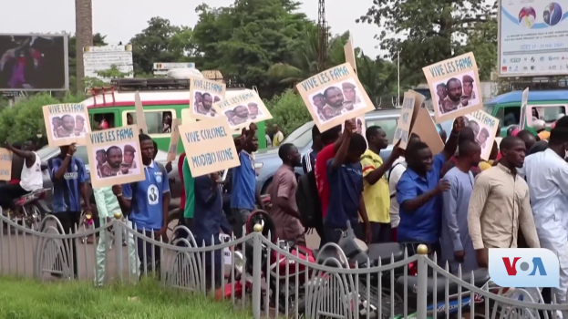 Eine Gruppe von Menschen mit Schildern steht vor einem Zaun, umgeben von Fahrzeugen, Bäumen, Pfählen, Werbetafeln und einem Bildschirm im Hintergrund, unter einem sichtbaren Himmel mit Gras am Boden des Bildes.