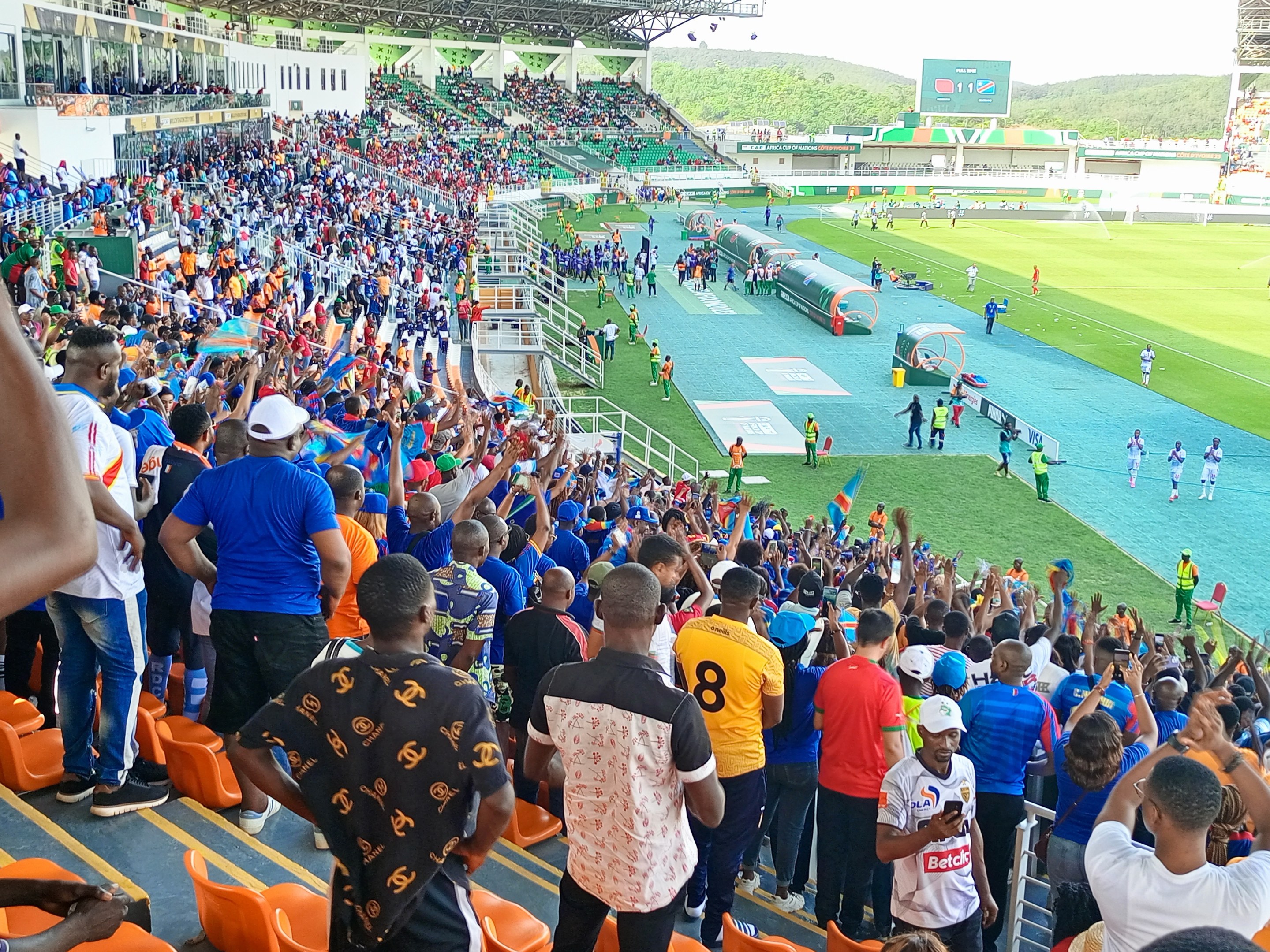 Eine große Menschenmenge versammelt sich vor einem eingezäunten Fußballfeld mit Hügeln und einem klaren blauen Himmel im Hintergrund, in Erwartung des Beginns eines Spiels während der 2019 African Cup of Nations.
