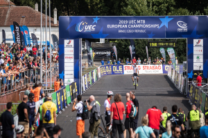 Radfahrer fahren unter einem Bogen mit der Aufschrift "2019 UEC MTB Europameisterschaften" eine Straße hinunter, gesäumt von Zuschauern und Bäumen, Gebäuden und einem klaren Himmel im Hintergrund.