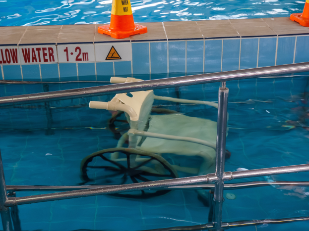 Schwimmbad mit einem Stuhl in der Mitte, umgeben von Sicherheitstonnen und einem Geländer sowie einem Schild an der Wand, das verfügbare Schwimmausrüstung anzeigt.