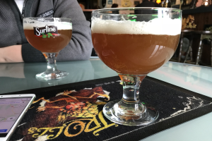 Zwei Gläser goldenes Bier auf einem Tisch mit einem Handy in der Nähe, umgeben von Menschen in einem Raum mit gerahmten Wanddekoration.