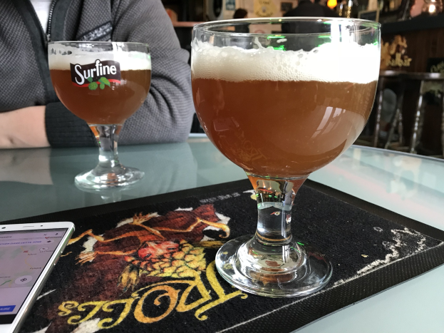 Zwei Gläser goldenes Bier auf einem Tisch mit einem Handy in der Nähe, umgeben von Menschen in einem Raum mit gerahmten Wanddekoration.