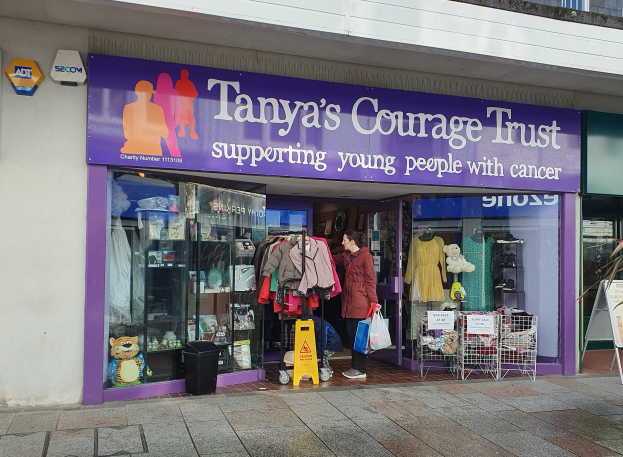 Ein Schaufenster mit einem Schild, das "Tanyas Mutvertrauen" trägt, eine Person davor und drinnen Kleidung, Teddybären, Puppen, ein Mülltonnen, ein Eimer, eine Pflanze und eine Tafel mit Text.