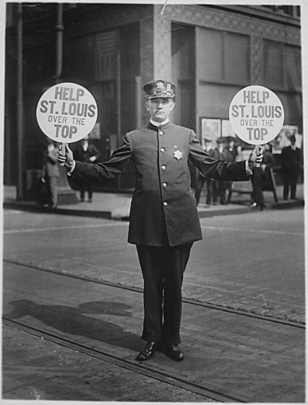 Ein altes Schwarz-Weiß-Foto eines Polizisten, der auf einer Straße steht und ein Schild hält, auf dem "Help St. Louis Over the Top" steht, mit ein paar Menschen und einem Gebäude im Hintergrund.