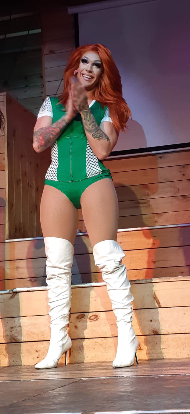 Eine Frau in einem grünen Bodysuit und weißen Stiefeln steht auf einer Bühne mit einer hölzernen Wand und einem Bildschirm im Hintergrund, bereit für eine Wrestling-Herausforderung.