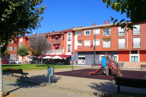 Ein Park mit einem Spielplatz, umgeben von Gebäuden, mit Bänken, Bäumen, Gras, einem Pfahl, einem Schild, einem Straßenpfahl, Fahrzeugen auf der Straße, Zelten, Menschen unter einem Dach sitzend und einem bewölkten Himmel.
