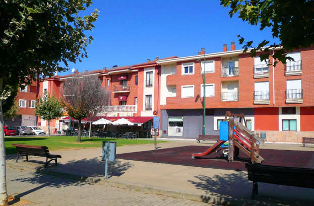Ein Park mit einem Spielplatz, umgeben von Gebäuden, mit Bänken, Bäumen, Gras, einem Pfahl, einem Schild, einem Straßenpfahl, Fahrzeugen auf der Straße, Zelten, Menschen unter einem Dach sitzend und einem bewölkten Himmel.