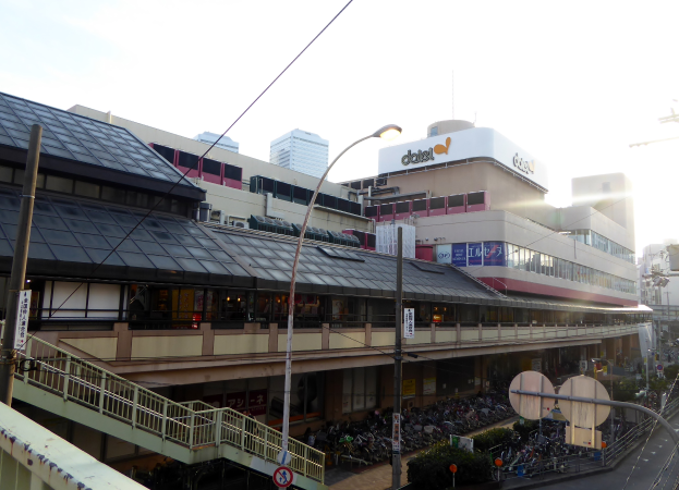 Eine belebte Stadtstraße mit starkem Verkehr, hohen Gebäuden, Straßenlaternen, Strommasten, Stromkabeln, Kraftfahrzeugen, Schildern, Büschen und einem bewölkten Himmel im Hintergrund.