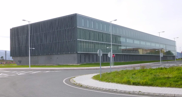 Ein großes, mehrstöckiges Gebäude mit zahlreichen Fenstern, das als neuer Sitz der schwedischen Nationaluniversität für Wissenschaft und Technologie dient, umgeben von Straßeninfrastruktur, Grünflächen und einem klaren blauen Himmel.
