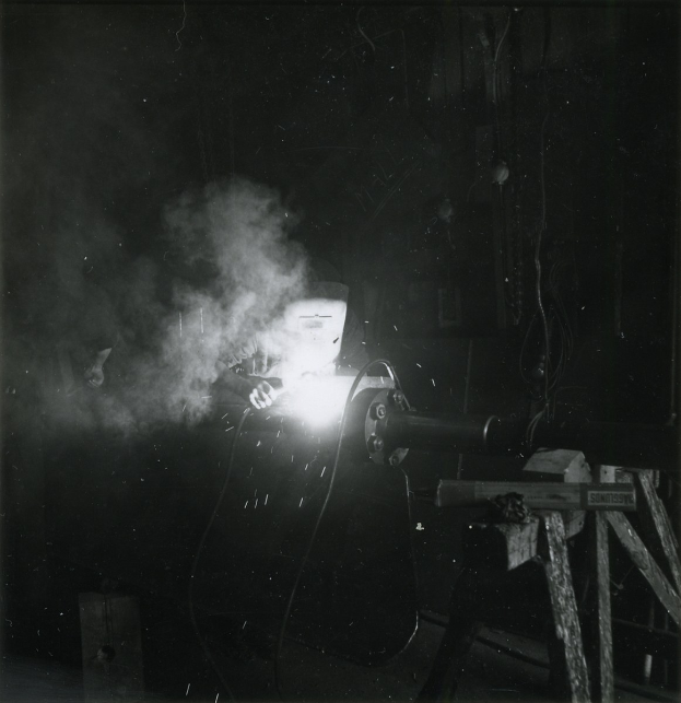 Schwarzes und weißes Foto eines Schweißers bei der Arbeit an Metall, mit Rauch von der Schweißmaschine und Hintergrundobjekten.