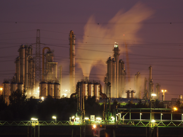 Ölraffinerie bei Nacht mit Rauchentwicklung, beleuchtet von Brücken-, Polen-, Baum- und Turmlichtern, vor einem sichtbaren Nachthimmel mit einem Wasserzeichen.