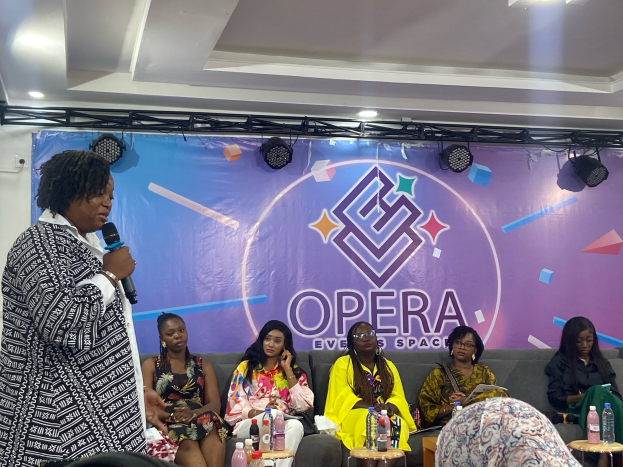 Eine Frau, die Gründerin des Opernveranstaltungsorts, steht mit einem Mikrofon vor einer Gruppe sitzender Frauen, mit Flaschen und anderen Gegenständen auf den Tischen im Vordergrund und einem Banner mit Text und Deckenbeleuchtung im Hintergrund.