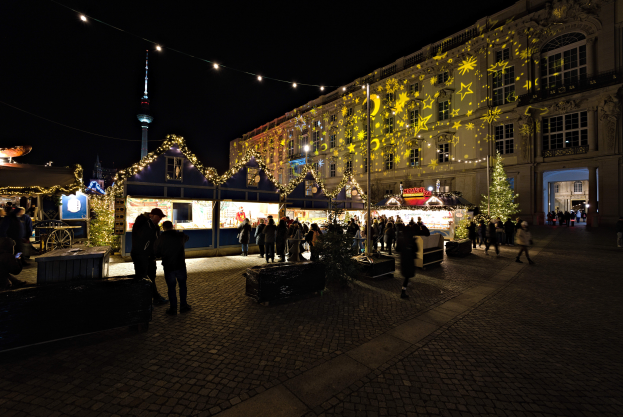 Ein lebendiger Weihnachtsmarkt in Berlin, Deutschland, mit Menschen um beleuchtete Stände, festliche Dekorationen und Gebäude mit beleuchteten Fenstern unter einem dunklen Himmel.