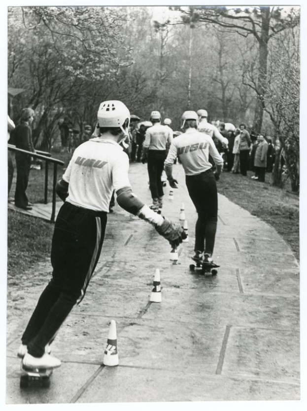 Eine Gruppe von Menschen in Helmen fährt auf einem Gehweg mit Verkehrskegeln, umgeben von Bäumen und einem Geländer, dargestellt in Schwarz-Weiß.