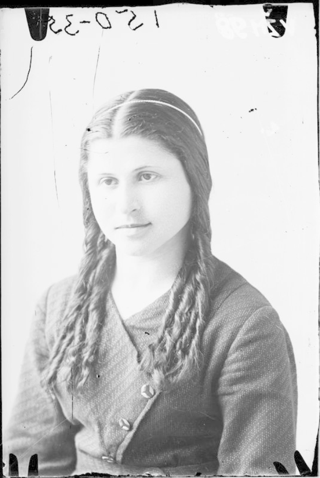 Ein Schwarz-Weiß-Porträt von Julia Margaret Cameron, eine Frau mit langen Zöpfen, die ein weißes Kleid und einen gemusterten Schal trägt und einen sanften Ausdruck hat, während sie ihre Hände vor sich verschränkt.