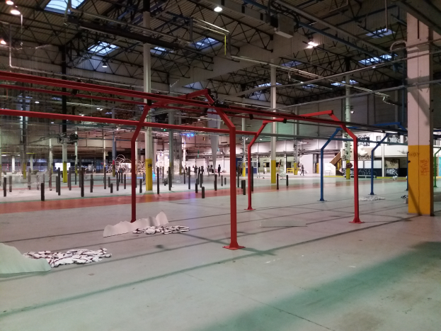Ein geräumiges Lagerinnere voller verschiedener Ausrüstungsgegenstände wie Stangen, Stäbe und Lichter, die auf dem Boden verstreut sind, mit ein paar Menschen im Hintergrund, was auf einen Indoor-Trampolinpark hindeutet.