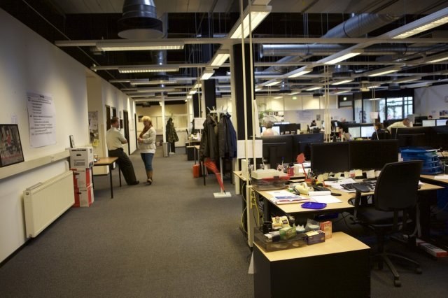 Großes Großraumbüro mit Menschen an Schreibtischen, Computern, Monitoren und anderen Büromöbeln; Wände zeigen Tafeln mit Text und Bildern, Deckenleuchten sind sichtbar und Glasfenster bilden den Hintergrund.