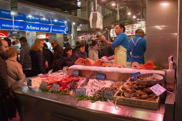 Ein Marktplatz mit Menschen, die gehen und einige Fische schneiden in der Mitte, mit Fischen und Brettern unten und Geschäften, Brettern, Pfählen und Lichtern im Hintergrund.