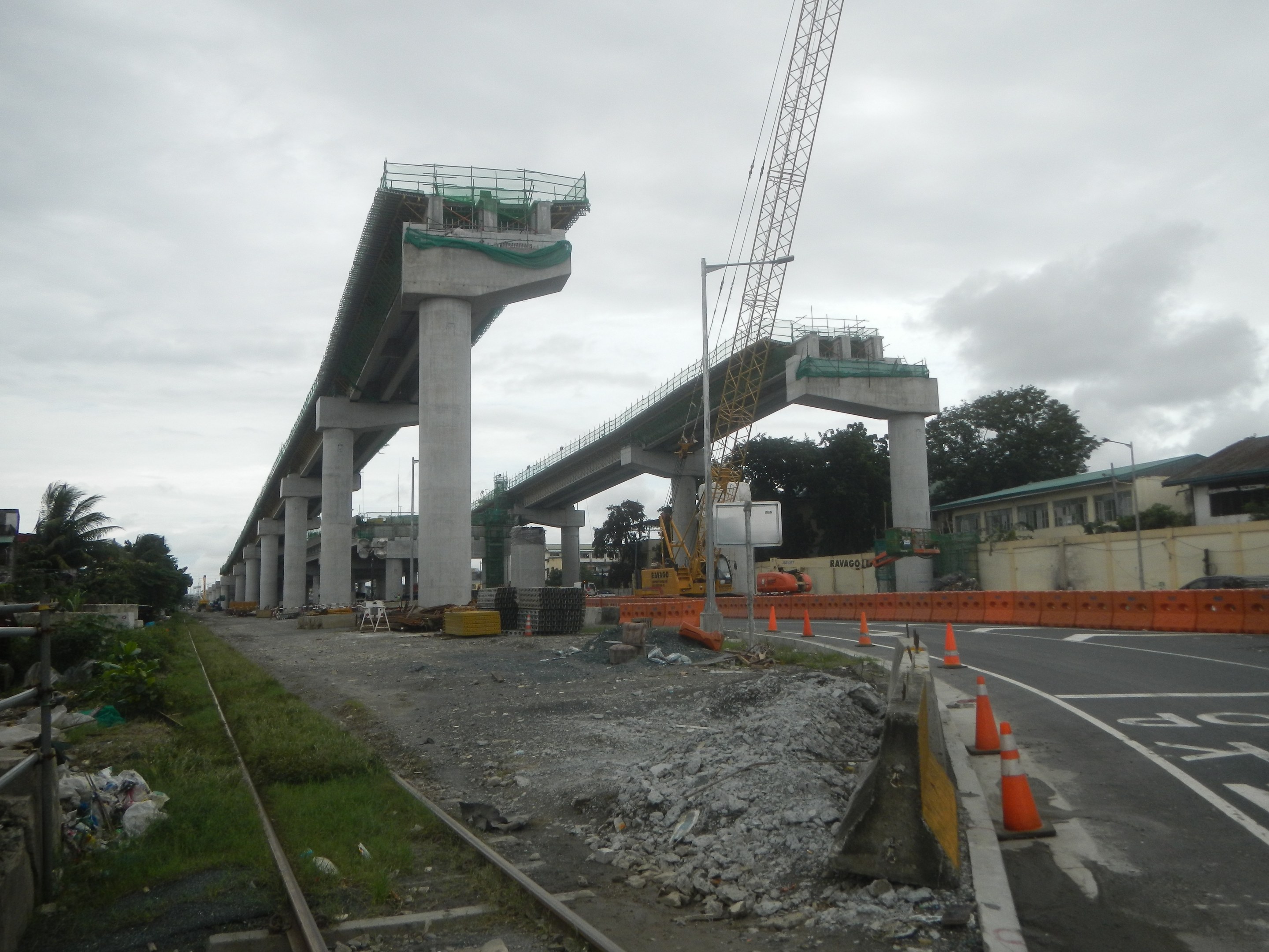 Baustelle mit einer Brücke im Hintergrund, Straße flankiert von Absperrbaken, verstreute Steine und Gras, Bahnschiene links, Bäume und Gebäude auf beiden Seiten und ein bewölkter Himmel.