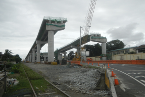Baustelle mit einer Brücke im Hintergrund, Straße flankiert von Absperrbaken, verstreute Steine und Gras, Bahnschiene links, Bäume und Gebäude auf beiden Seiten und ein bewölkter Himmel.