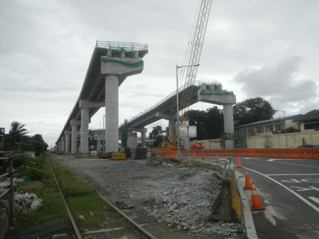 Baustelle mit einer Brücke im Hintergrund, Straße flankiert von Absperrbaken, verstreute Steine und Gras, Bahnschiene links, Bäume und Gebäude auf beiden Seiten und ein bewölkter Himmel.