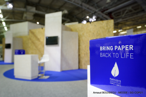 Ein Messestand mit einem blauen und weißen Schild, auf dem "Bring Paper Back to Life" steht, auf der rechten Seite, im Hintergrund sind leicht unscharfe Stühle, Tische, ein Fernseher und Deckenleuchten zu sehen.