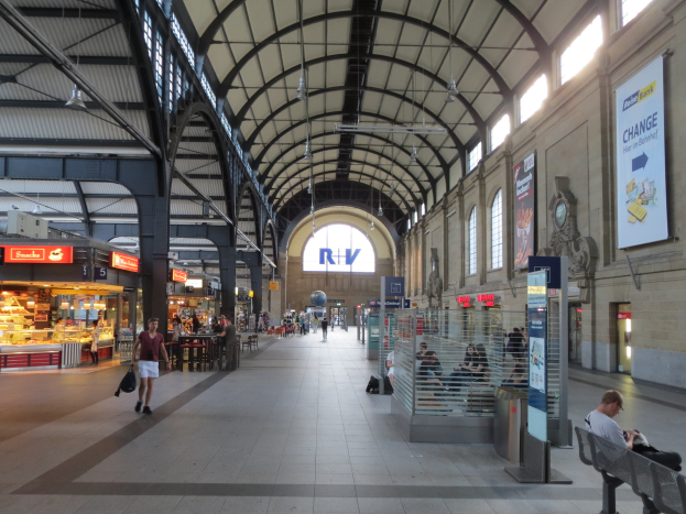 Ein belebter Bahnhof mit Menschen, die gehen, auf Bänken sitzen und stehen, umgeben von Texttafeln, Lampen und anderen Gegenständen, mit einer Uhr an der Wand im Hintergrund.