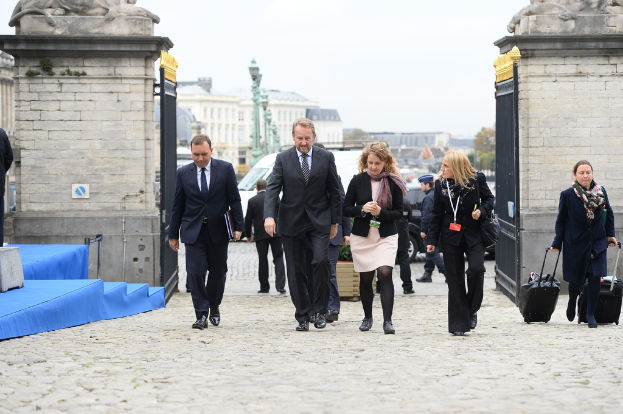Eine Gruppe von Mitarbeitern geht in einen Bereich mit einem Tor, jenseits dessen sich viele Fahrzeuge und Gebäude befinden; rechts trägt eine Frau zwei Gepäckstücke.