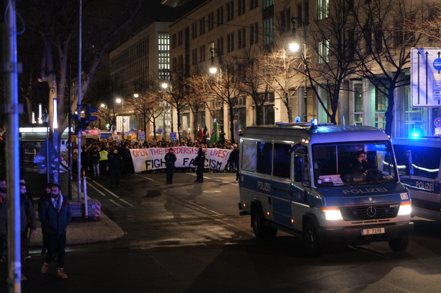 Eine Gruppe von Menschen, die nachts eine von Straßenlaternen beleuchtete Straße entlanggeht, mit einem Polizeiwagen im Vordergrund und einem Banner mit Text im Hintergrund.