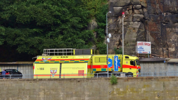 Ambulanz auf der Straße neben einem Felsen geparkt, mit verschiedenen Straßenobjekten im Vordergrund und Bäumen und einem Auto im Hintergrund.
