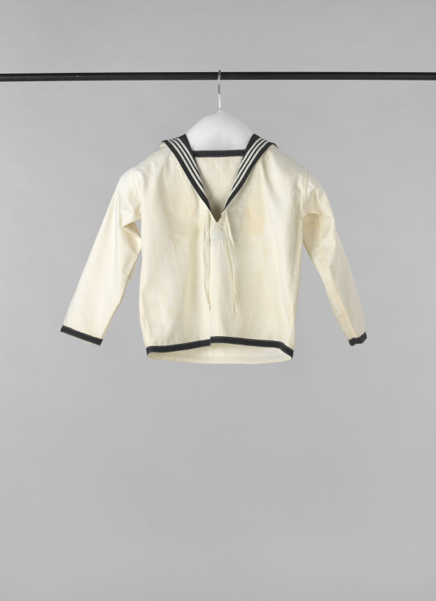 Eine weiße Matrosenjacke mit Kragen, langen Ärmeln und Knopfleiste hängt an einer Wäscheleine vor einem weißen Hintergrund, aus leichtem, geripptem Stoff hergestellt.