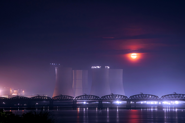 Ein Kernkraftwerk bei Nacht, mit einer Brücke über einen Gewässser, beleuchtet von Gebäudelichtern und dem Mond, umgeben von Bäumen.