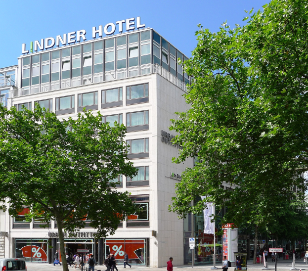 Das Lindner Hotel in Berlin, ein großes Glasfenstergebäude mit einer Aufschrift auf dem Dach, umgeben von Bäumen und einer belebten Straße mit Füßgängern und Fahrzeugen.