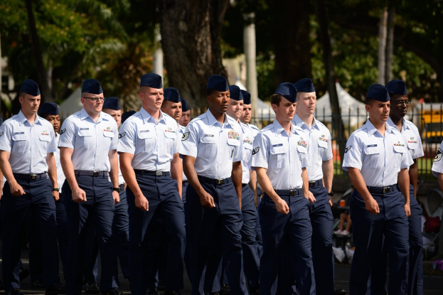 M√§nner in Uniform auf einer Stra√Ÿe mit einem Eisenzaun dahinter, B√§ume und Masten im Hintergrund.
