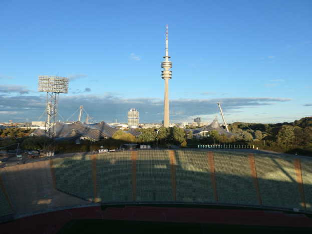 Olympiastadion in Berlin, Deutschland, mit dem Fernsehturm (Fernsehturm) im bewölkten Hintergrund, umgeben von Bäumen, Gebäuden und Lichtern.