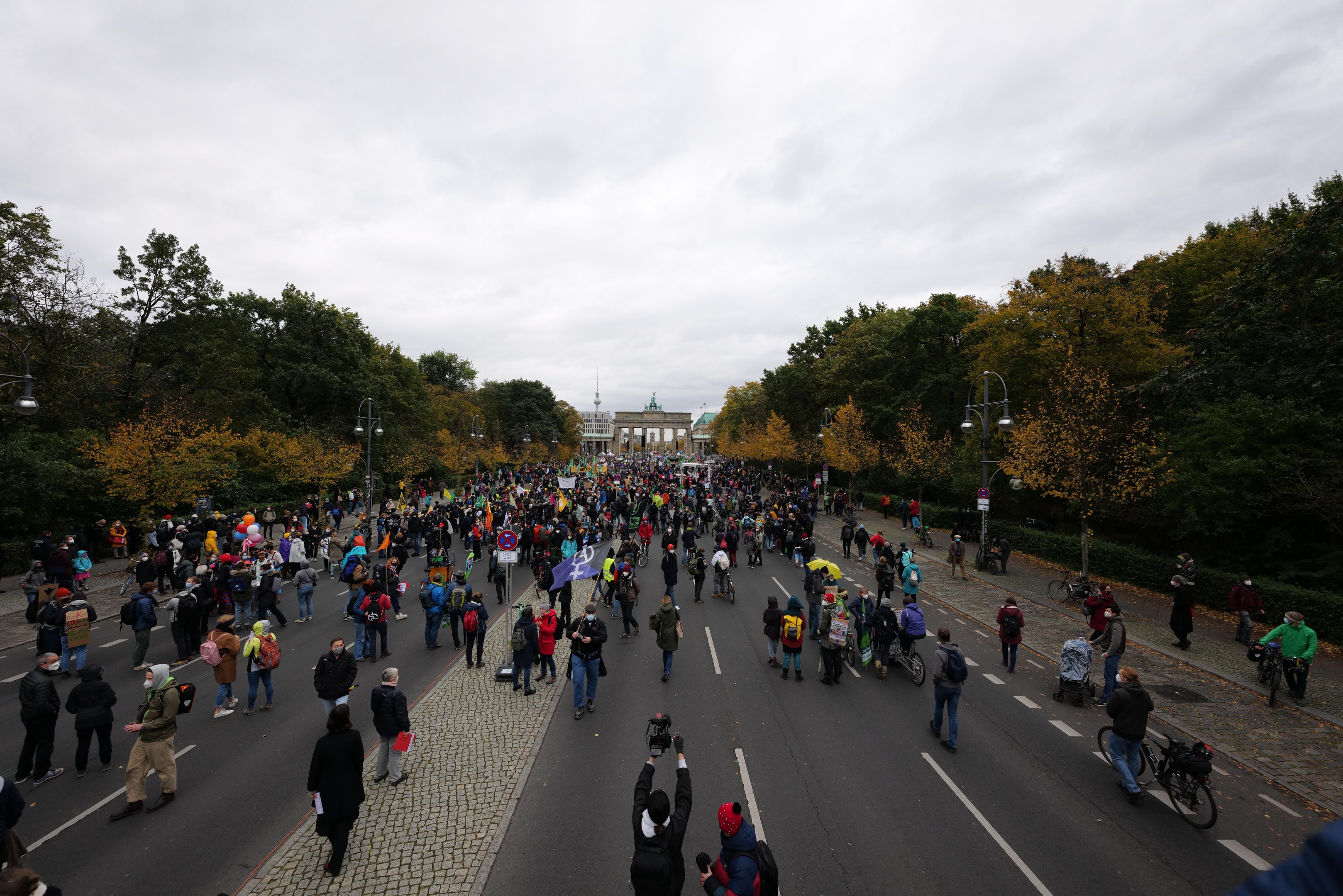 Eine große Gruppe von Menschen marschiert auf einer von Bäumen gesäumten Straße in Berlin, mit Kameras bewaffnet, mit einem Gebäude und einem klaren Himmel im Hintergrund.