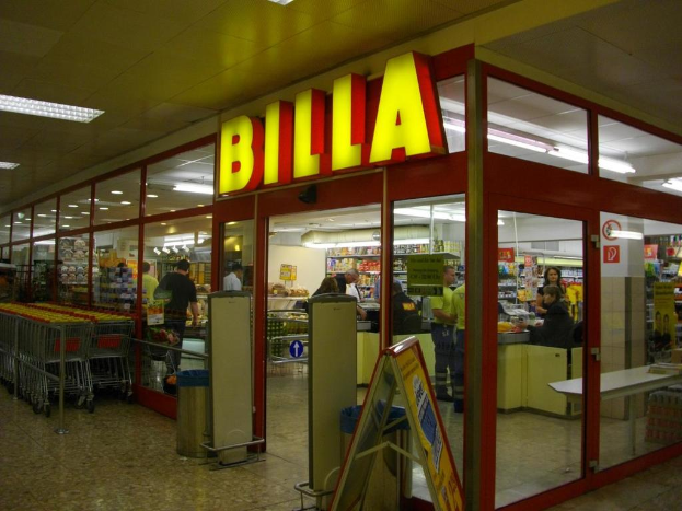 Ein Supermarkt mit einem Eingang in der Mitte, Mülltonnen daneben, eine Reihe von Einkaufswagen auf der linken Seite und ein paar Menschen drinnen, die unter Deckenbeleuchtung Produkte durchstöbern.