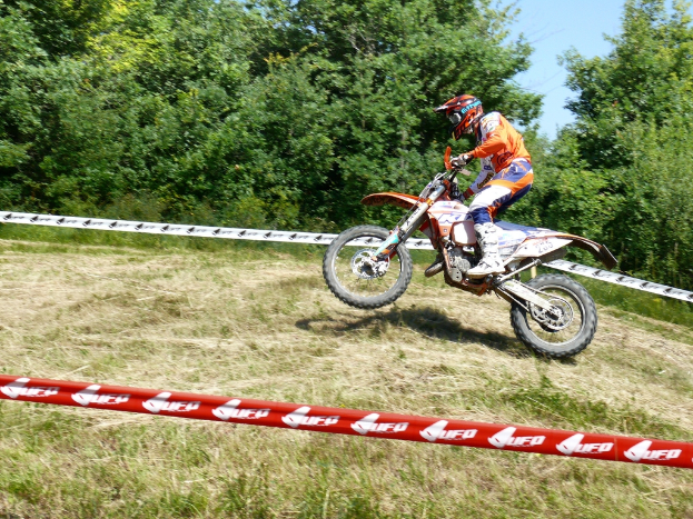Eine Person mit Helm fährt ein Fahrrad umgeben von Absperrband, mit Bäumen, Gras und Himmel im Hintergrund.