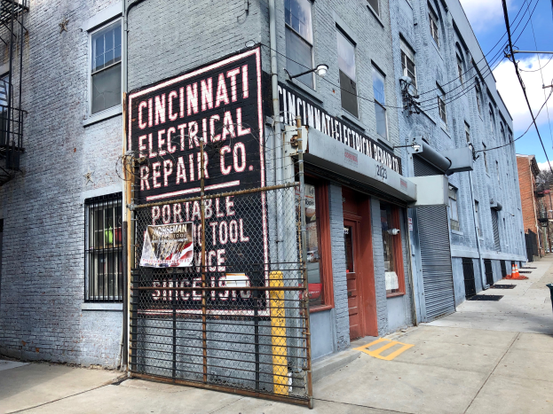 Das Cincinnati Electrical Repair Co. Gebäude mit einer Werbetafel, Metallzaun, Mülltonne, Pfählen, Drähten, Bäumen und einem bewölkten Himmel.