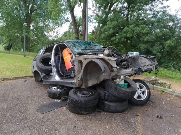 Ein umgestürzter Wagen am Straßenrand umgeben von Trümmern, Gras, Pflanzen, Bäumen, einem Laternenmast und einem bewölkten Himmel.