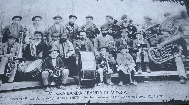 Ein Schwarz-Weiß-Foto einer Band von Musikern, die posen, einige sitzen und einige stehen, halten verschiedene Musikinstrumente und Text am unteren Rand des Bildes.