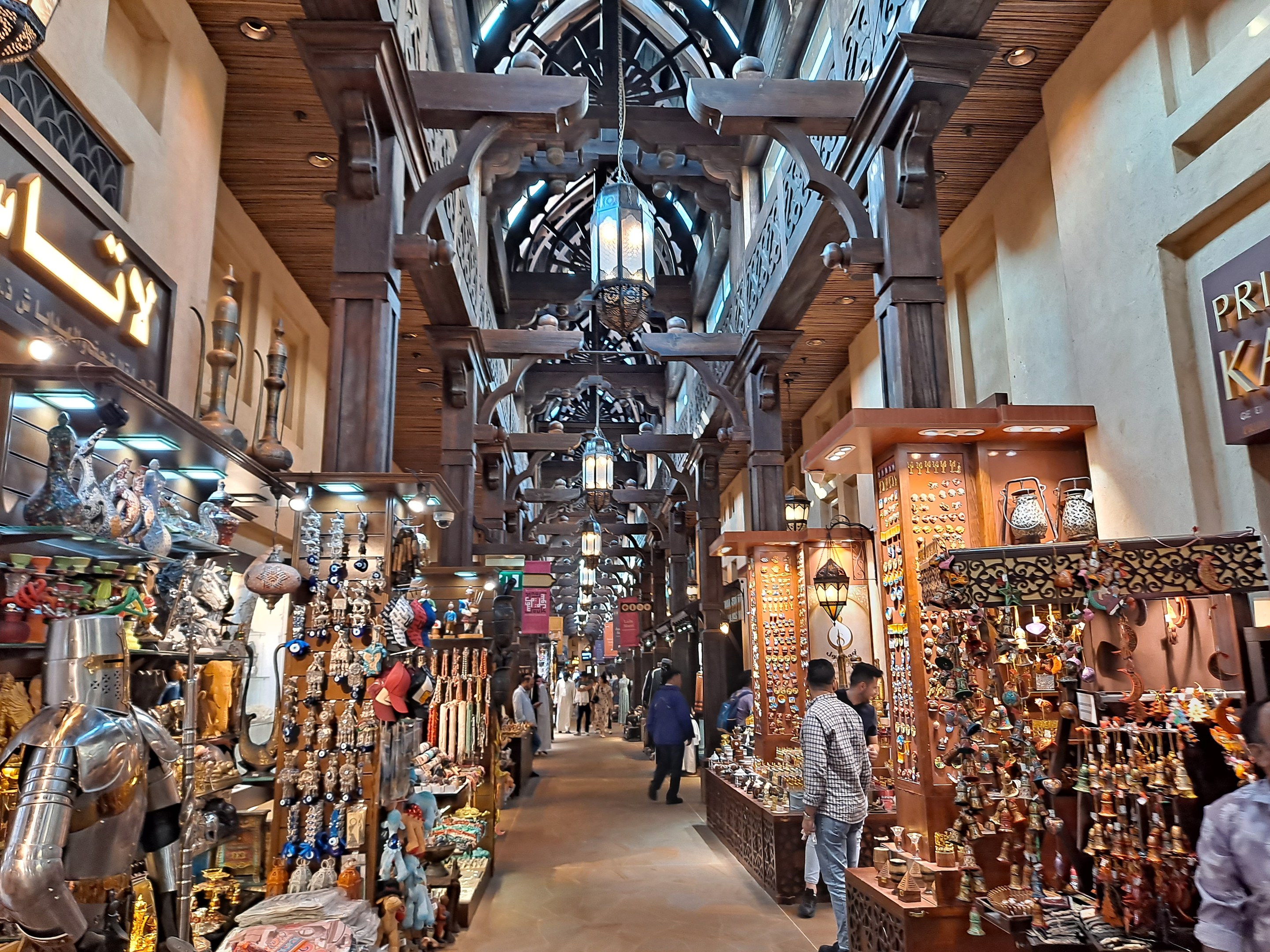 Ein geschäftiges Dubai-Bazar mit Menschen, die zwischen Regalen voller Waren unter einer mit warmem Licht beleuchteten Decke hindurchgehen, und Texttafeln an den Wänden.