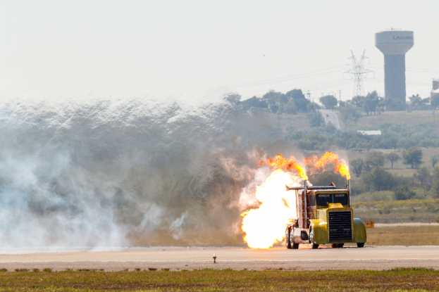 Ein gelber Traktor fährt auf einer Straße mit großen Flammen, umgeben von Gras, mit Bäumen, Türmen, Drähten und einem klaren blauen Himmel im Hintergrund.