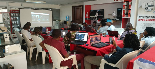 Eine Gruppe von Menschen sitzt um einen Tisch mit Laptops, Mäusen und Flaschen, nimmt an einem digitalen Marketing-Workshop teil, mit einem Banner, einem Display-Bildschirm und elektronischen Geräten im Hintergrund.