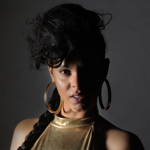 Eine Frau in einem goldenen Kleid und großen Ohrringen steht selbstbewusst vor einer Wand, ihre glatten Bob-Frisur und das natürliche Make-up betont durch pinken Rouge und glänzende Lippen, sieht direkt in die Kamera mit einem entschlossenen Ausdruck.