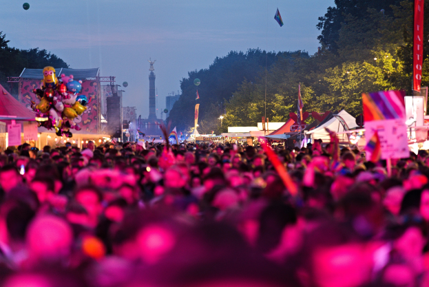 Eine große Menge bei einem nächtlichen Musikfestival, beleuchtet von bunten Lichtern, mit Zelten, Fahnen und Pfählen im Vordergrund, Bäumen und einem Turm im Hintergrund und einem klaren Nachthimmel.