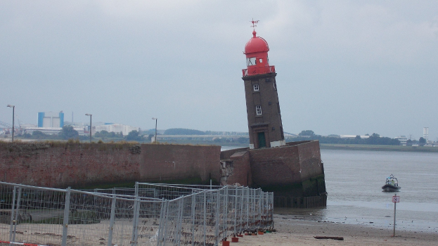 Roter Leuchtturm auf einem sandigen Strand neben Wasser mit einem Boot, einer Wand links, Bäumen und Gebäuden im Hintergrund unter einem klaren blauen Himmel.