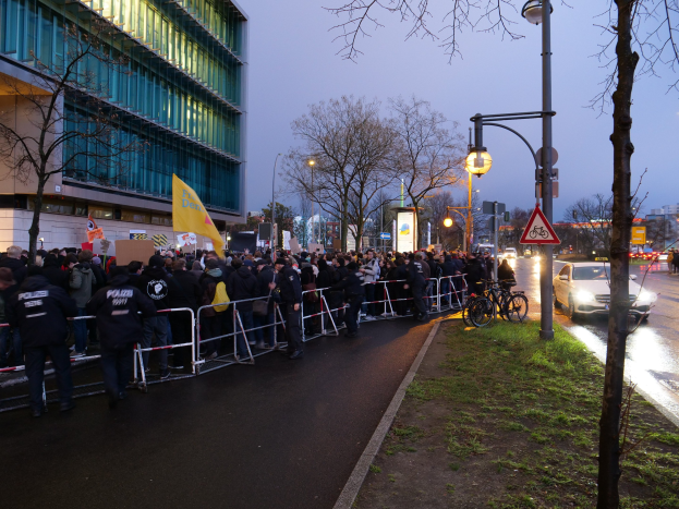 Eine große Gruppe von Menschen mit Schildern steht vor einem Gebäude mit Barrikaden und Bäumen, was auf eine Protestaktion in Berlin hinweist.