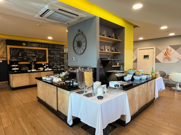 Ein Restaurantinnenraum mit Tischen und Stühlen, Speisen, einer Tafel mit Text, einer Wanduhr, Deckenleuchten, einer Klimaanlage und einer Tür im Hintergrund, gelegen im Hôtel de Ville in Paris.